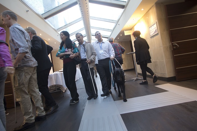 Sehende und sehbehinderte Delegierte gehen mit Führhunden und weißen Stöcken, den Leitstreifen nutzend, auf dem Hotelflur entlang