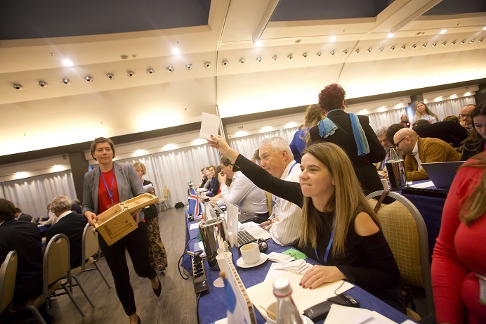 A female delegate raise her hand with a voting slip, showing readiness to vote, as an assistant approaches with a ballot box.