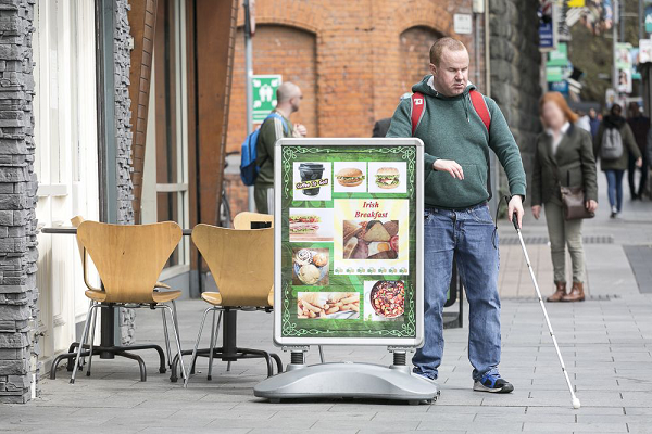 A blind man with a white cane attempting to navigate around a shop sign in the street.