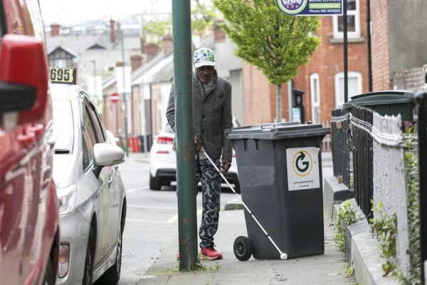 A man with a white cane trying to navigate between a dustbin and a lamppost on the street.