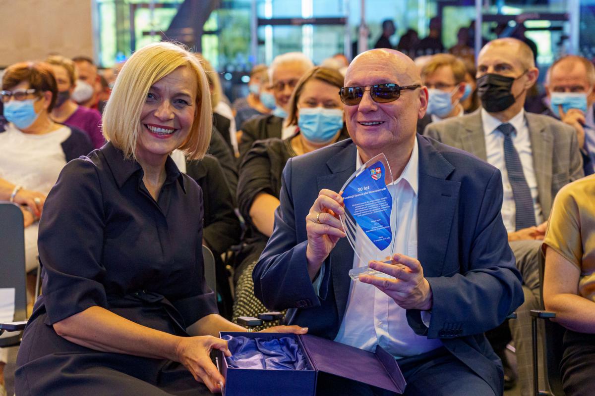 the smiling honorary president of our organization with the statuette - 30 years of the Foundation, sitting among the participants of the REHA conference. People in the background are wearing masks. The background is slightly blurred.